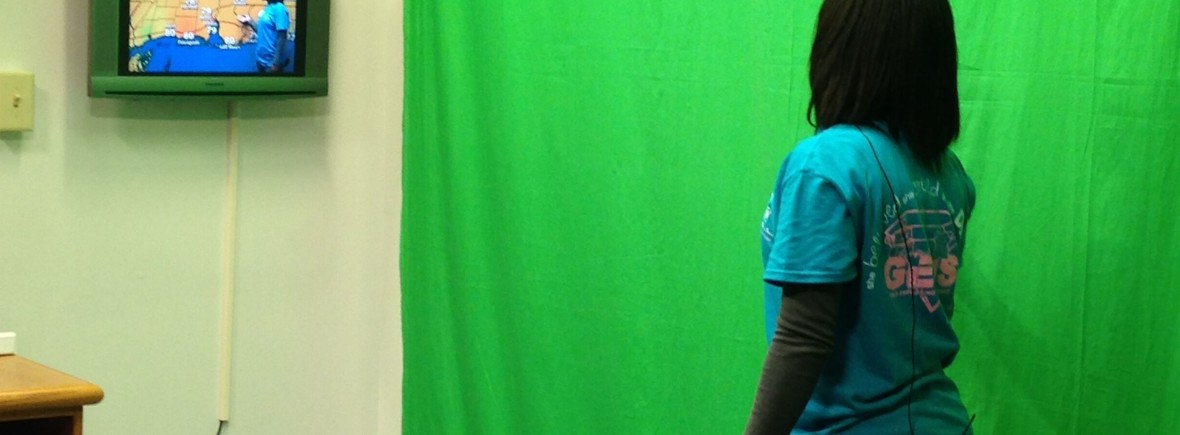 A student stands in front of a green screen and sees herself forecasting the weather on a television.